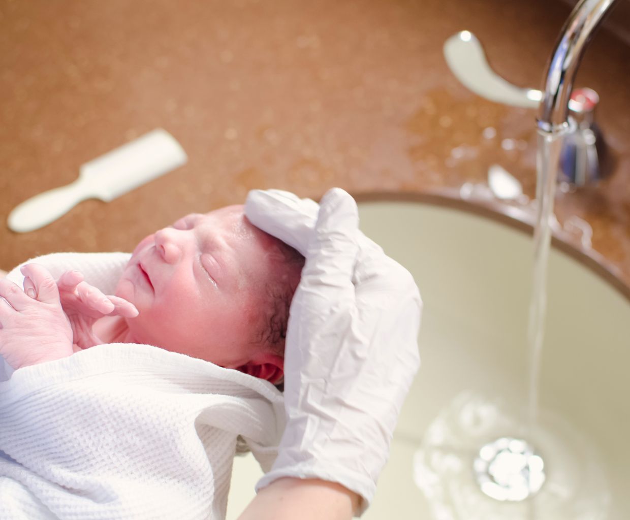 Newborn wrapped in a towel having their head gently washed with running water at a sink