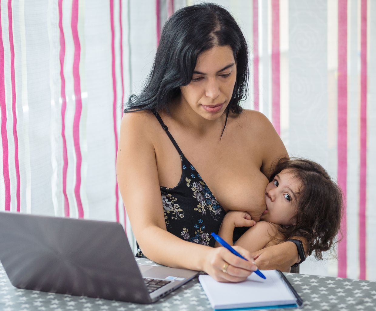 Mother writing in a notebook at a table while breastfeeding her toddler, who looks at the camera