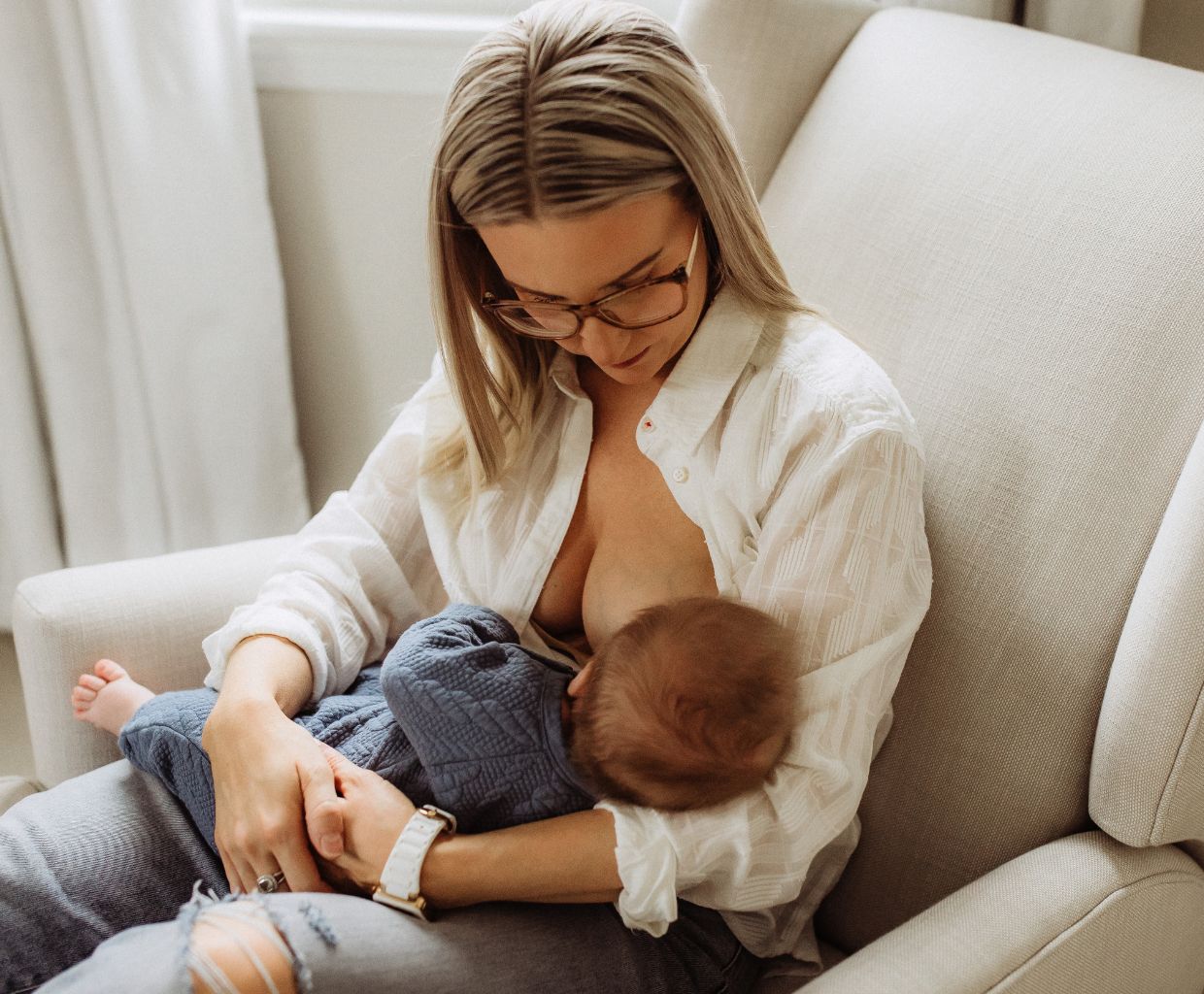 Mother in glasses and a white shirt breastfeeding her baby in a cream armchair