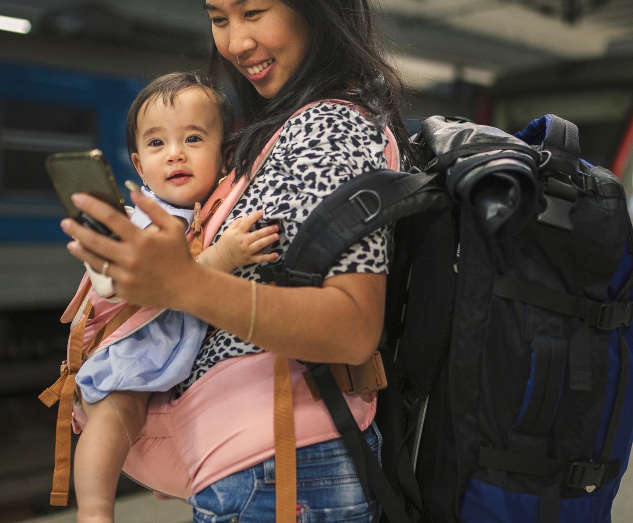 Smiling mother carrying her baby in a front carrier and a large backpack at a train station