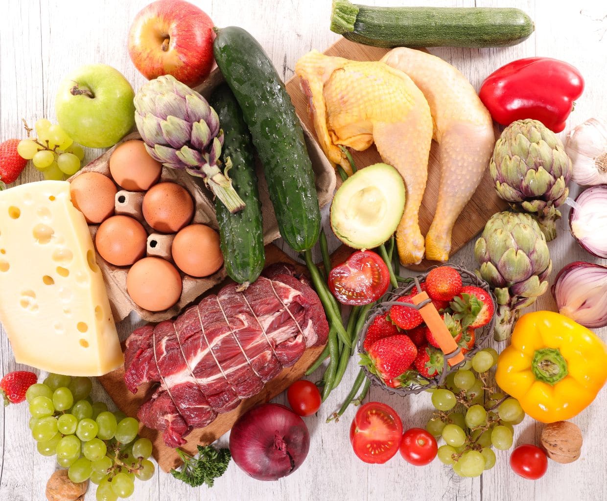 Fresh vegetables, fruit, eggs, cheese, chicken and beef laid out on a white wooden table