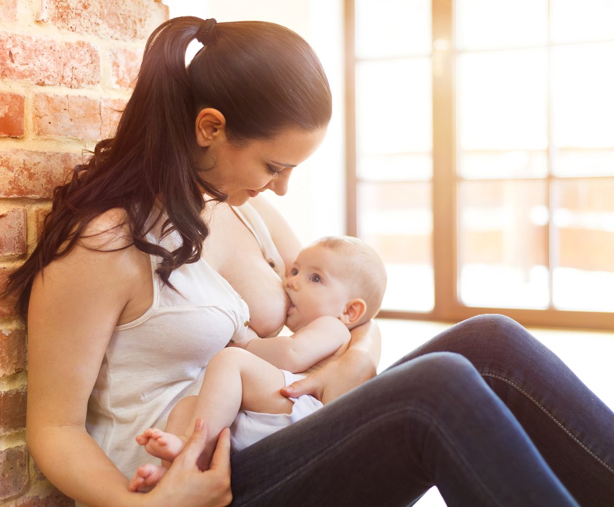 Smiling mother sitting by a brick wall breastfeeding her baby in a cradle hold