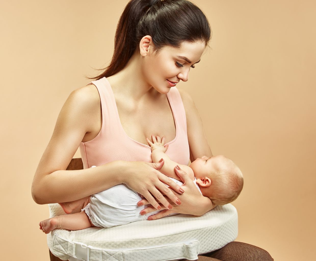 Mother sitting upright and smiling at her baby, who lies across a firm white nursing pillow on her lap