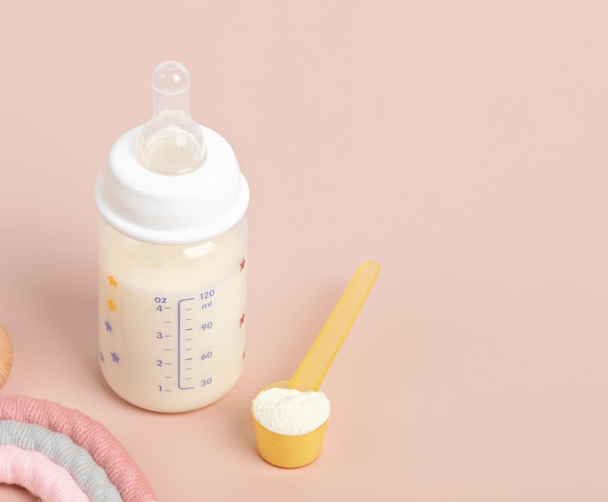 Baby bottle of prepared formula next to a yellow measuring scoop of formula powder on a pink background