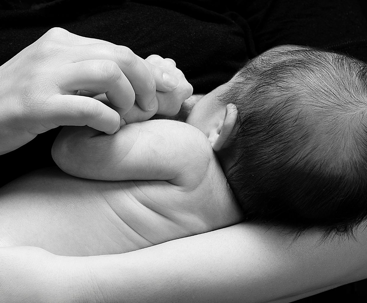 Black-and-white photo of a newborn breastfeeding while holding a parent's finger