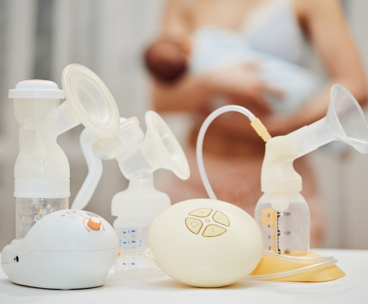 Several electric and manual breast pumps on a table, with a mother holding her baby in the background