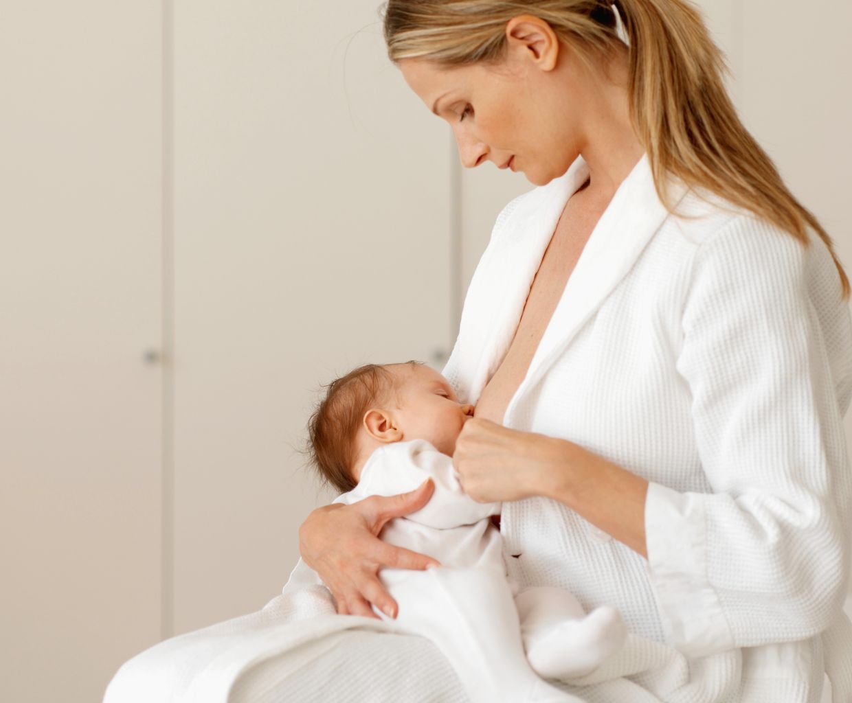 Mother in a white robe calmly breastfeeding her newborn while seated