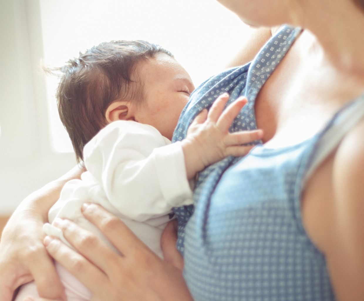 Sleepy newborn in a white bodysuit breastfeeding in a parent's arms in soft daylight