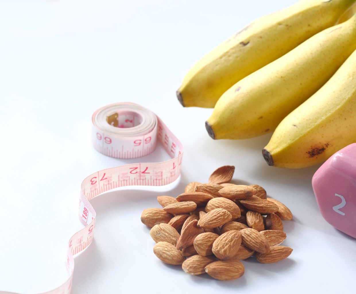 Tape measure, almonds, bananas and a small pink dumbbell on a white table