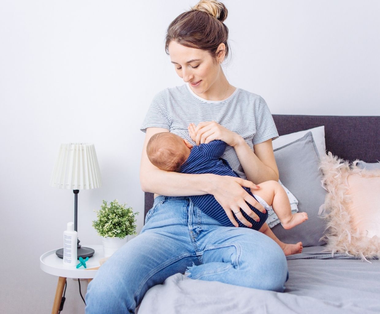 Smiling mother sitting on a bed breastfeeding her baby, with a lamp and plant on the nightstand