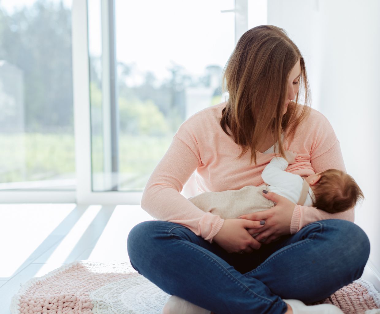 Mother sitting cross-legged on a rug by a window, breastfeeding her baby in the cradle hold
