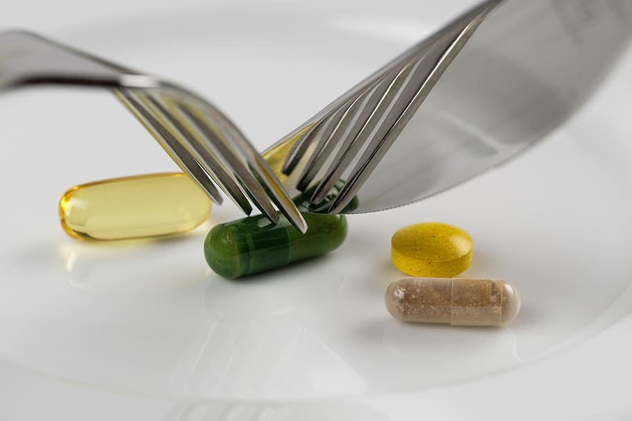 Assorted supplement capsules and tablets on a white plate beside a fork and knife