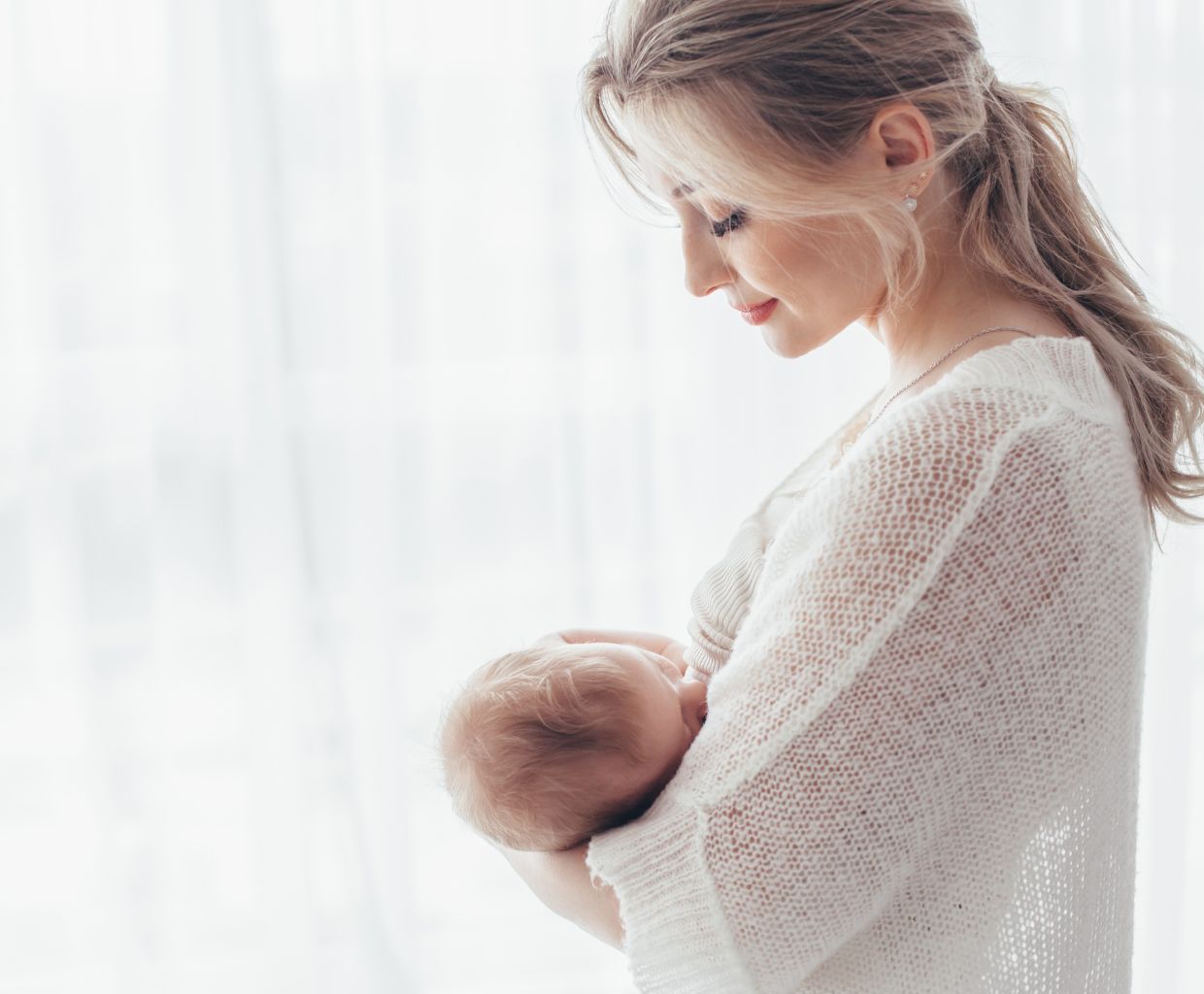 Mother in a white knit sweater breastfeeding her baby by a bright window