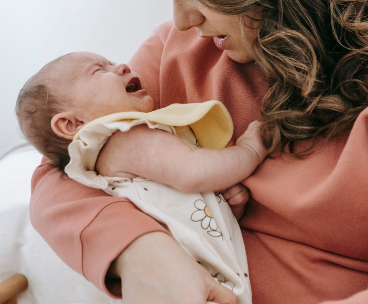 Mother in a coral sweater holding her crying baby close and looking down at them