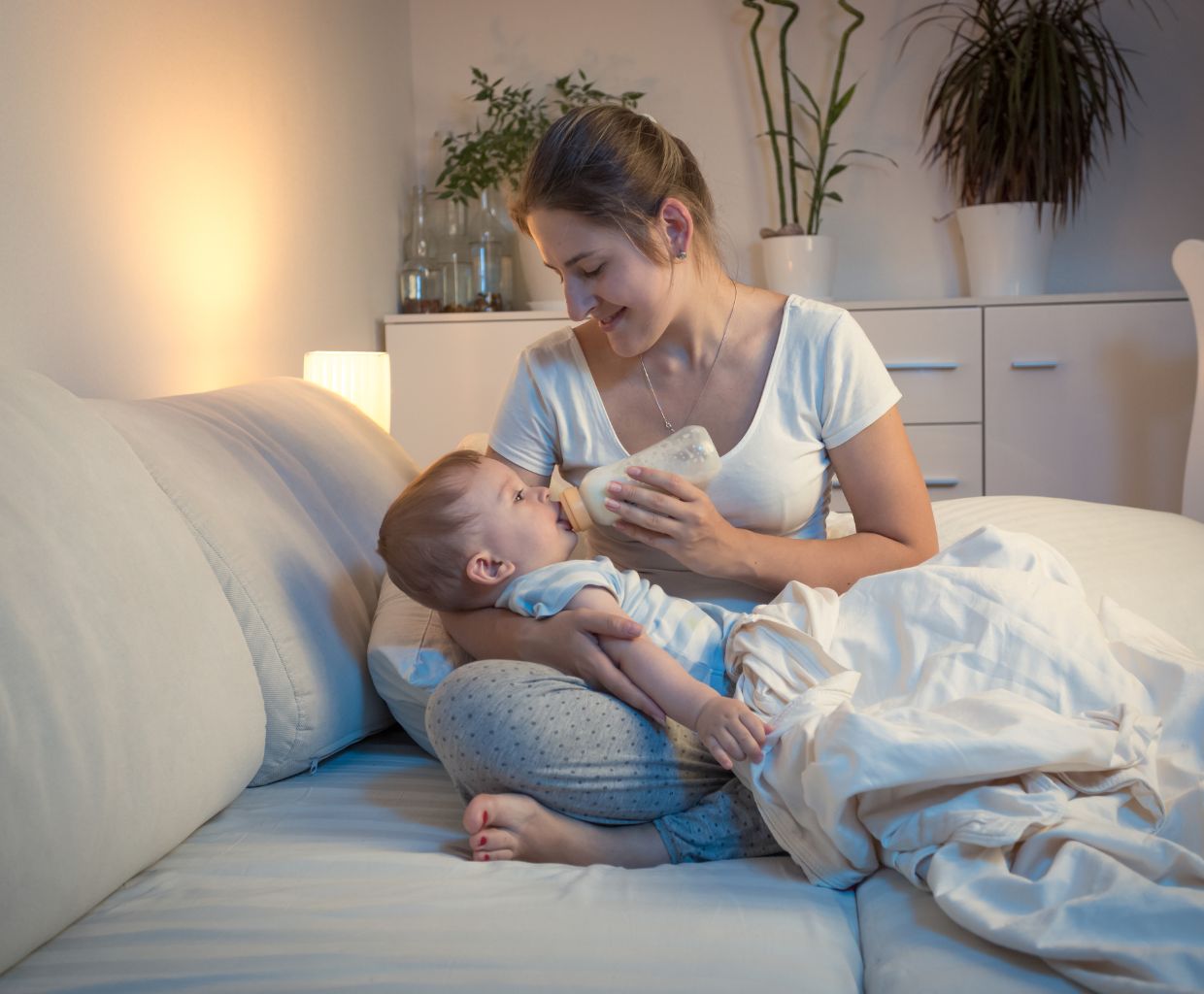 Mother smiling at her baby while giving him a bottle on a bed in a softly lit bedroom
