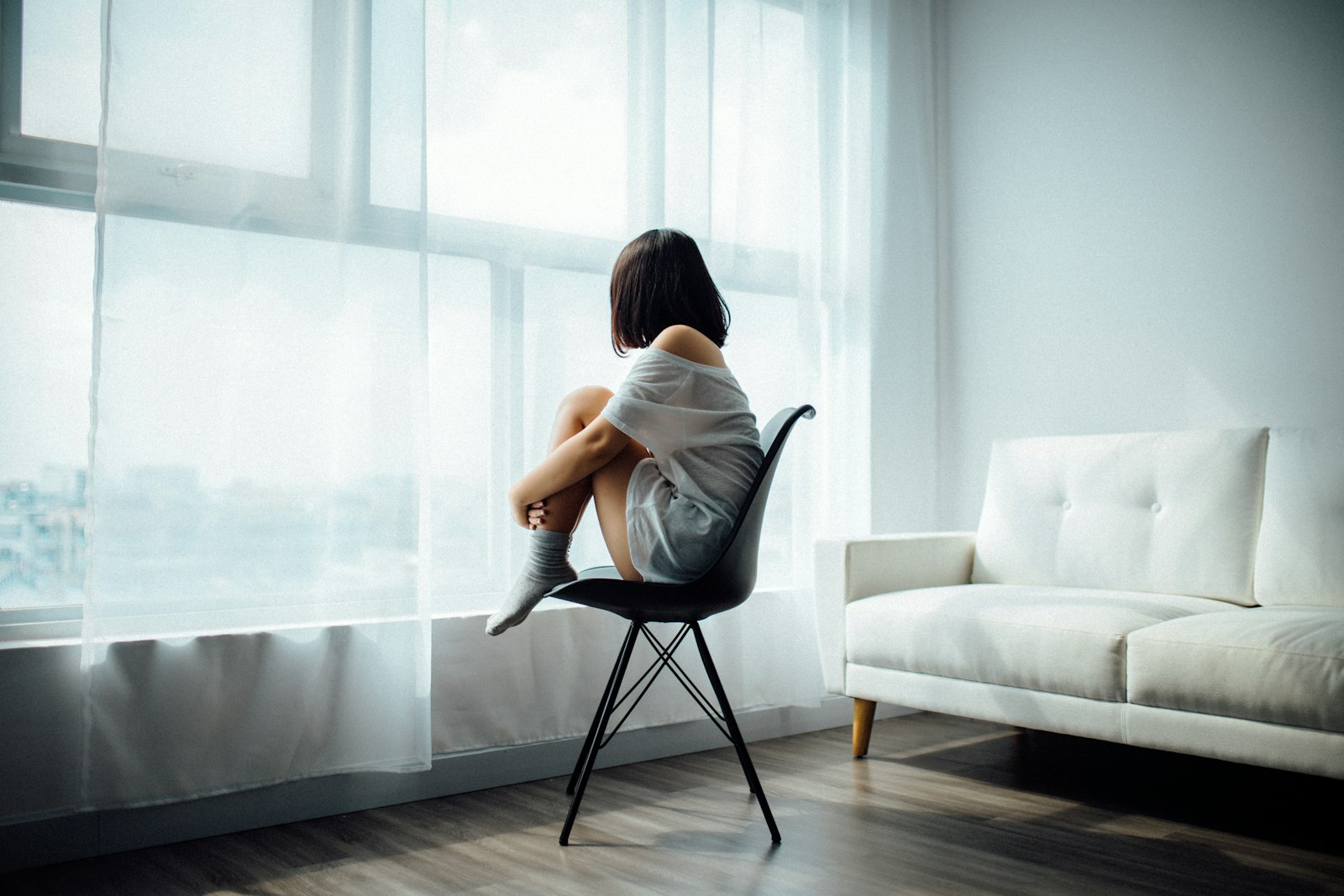 Woman sitting alone on a chair by a bright window, hugging her knees