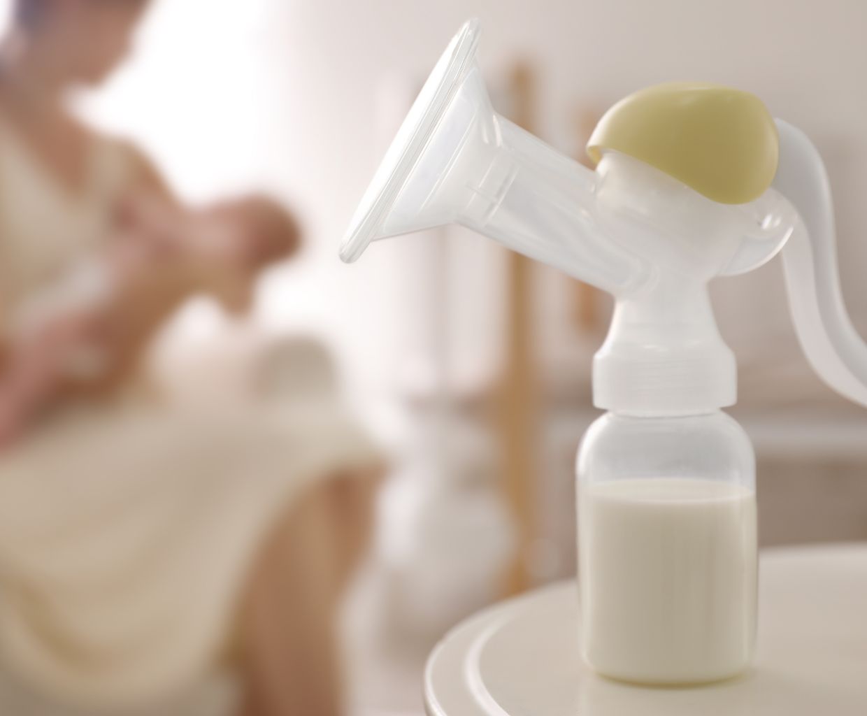 Manual breast pump with a bottle of expressed milk on a table, a mother holding her baby in the background