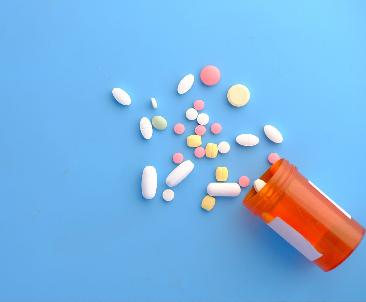 Assorted white, pink and yellow tablets spilling from an orange pill bottle on a blue background