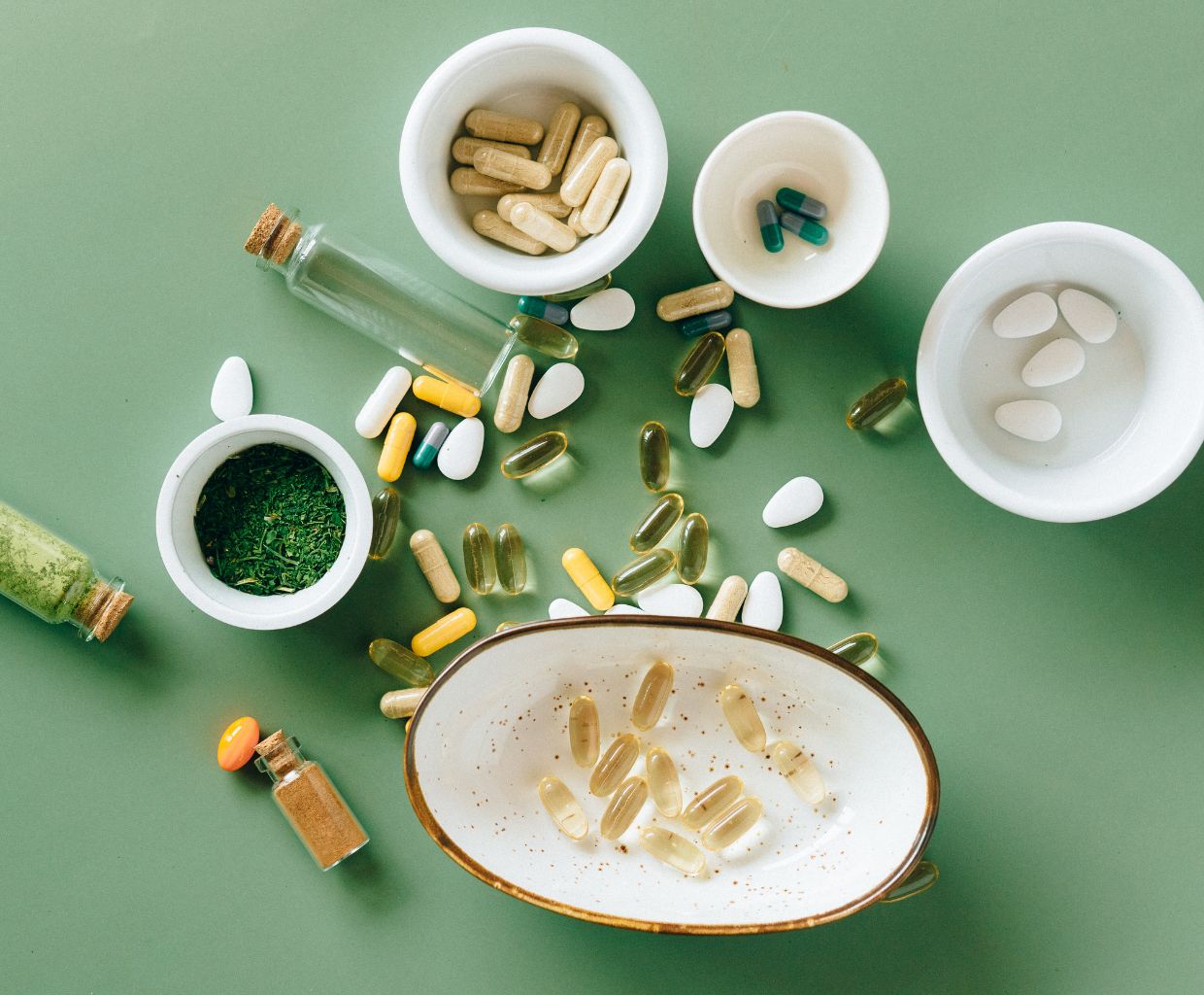 Assorted supplement capsules and softgels in white bowls on a green background
