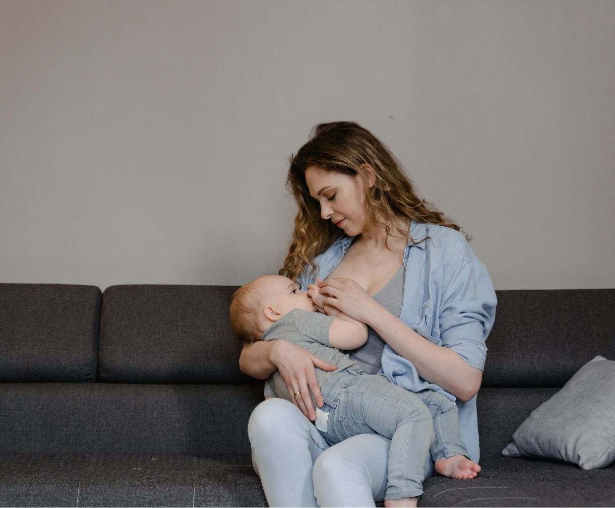 Mother sitting on a gray sofa breastfeeding her older baby and looking down at him