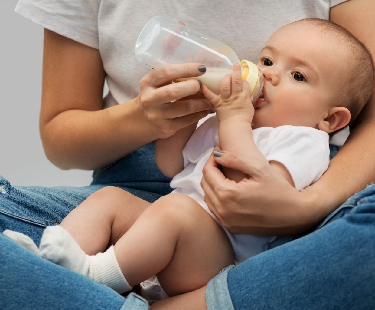 Baby in a white onesie drinking expressed milk from a bottle held by a parent
